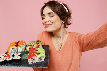 Close up young woman in sweater hold in hand sniff makizushi sushi roll served on black plate traditional japanese food doing selfie shot pov on mobile phone isolated on plain pastel pink background