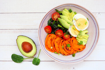 Breakfast nutrient bowl with sweet potato, egg, avocado and spinach. Top view table scene on a white wood background.