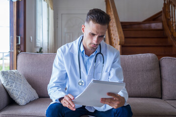 Portrait of handsome young male doctor in white lab coat with stethoscope looking at reports in...