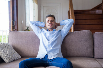 Satisfied young man relaxing while sitting on couch with hands behind head at home. Handsome...