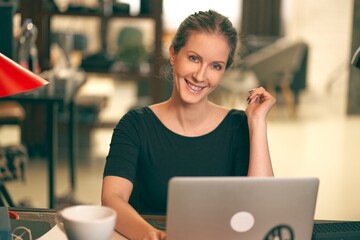 Portrait of young brown hair business woman sitting at desk table in the office, working at home, using laptop computer, looking at camera.