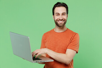 Young smiling happy cheerful man 20s wearing casual orange t-shirt hold use work on laptop pc computer isolated on plain pastel light green color background studio portrait. People lifestyle concept.
