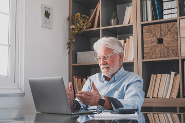 Senior businessman video chatting online using laptop at home office in front of book shelf. Elderly caucasian man gesturing while working online or watching media content at workplace