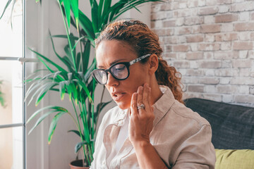 Young woman suffering from tooth pain and touching cheek while sitting on couch at home. Caucasian...