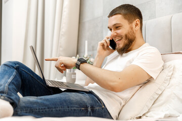 young man at home on the couch talking on the phone and looking at the laptop.