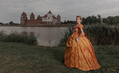 A young woman in an old orange dress walks on the shore of the lake near the castle