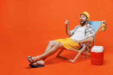Young amazed tourist man in beach shirt hat lie on deckchair near fridge point finger aside on...