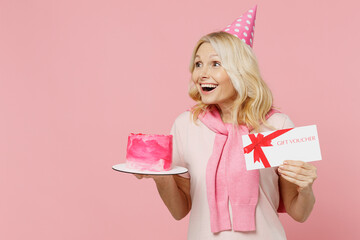 Elderly amazed woman 50s in t-shirt birthday hat hold cake gift certificate coupon voucher card for store look aside isolated on plain pastel pink background studio Celebration party holiday concept