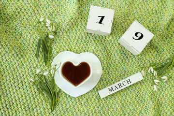 Calendar for March 19: cubes with the number 19, the name March in English, a cup of tea in the shape of a heart, snowdrops on a knitted knitted fabric, top view