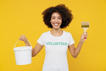 Young woman of African American ethnicity wears white volunteer t-shirt hold paint bucket brush working isolated on plain yellow background. Voluntary free work assistance help charity grace concept. © ViDi Studio