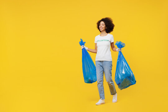Full body young woman of African American ethnicity in white volunteer t-shirt carry trash bags after rubbish removing isolated on plain yellow background. Voluntary free work assistance help concept.