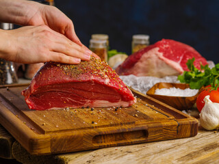 The chef rubs spices on a large piece of raw meat - pork, beef on a wooden cutting board. Close-up....