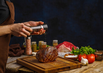 Cooking a festive pork ham by the hands of a professional chef on a wooden cutting board on a dark blue background. Meat, herbs, vegetables, spices in the background. Restaurant cuisine recipes.