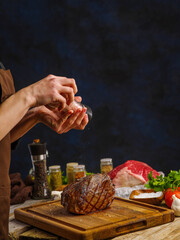 The chef cooks a pork ham on a dark blue background. Lots of vegetables, spices, herbs. Restaurant, hotel. Recipes for restaurant and home cooking, a festive meat dish.