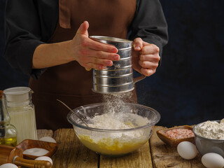 A professional chef in a dark uniform sifts flour on a wooden table, dark blue background. Close-up. Hotel, restaurant, confectionery, home cooking. Advertising business, banner.