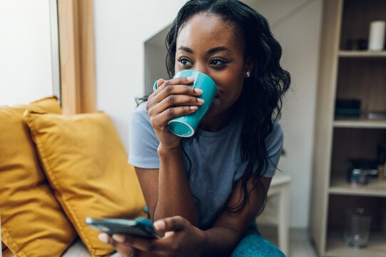 African american woman sitting on windowsill with a cup of coffee and a smartphone