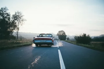 Fotobehang A retro car drives along the road at dawn. The convertible drives off into the distance on a deserted road © Anna