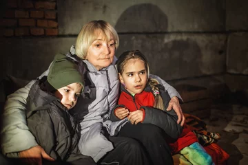 Zelfklevend Fotobehang Grandmother hugs her little kids while they sit out the attack of the Russian aggressor in a bomb shelter. Terrorism and war, current Ukrainian and world history © Maria