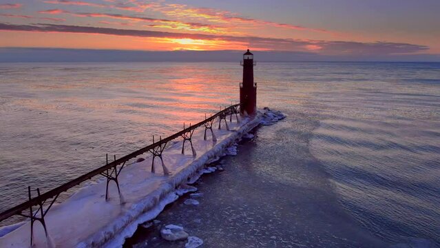 There is amazing beauty in this aerial view of an icy harbor in Winter, clouds on the horizon, waves reflecting the fiery sky, lighthouse beacon blinking, Lake Michigan in the Winter just before sunri