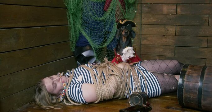 Tied with ropes woman captured by pirate tries to break free lying on wooden floor near dressed dachshund IN hat against green mesh
