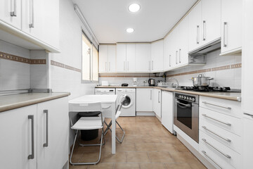 Kitchen with a lot of white furniture, a white square table with folding chairs and various accessories on the counter