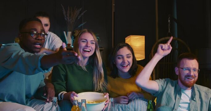 Excited young multiethnic friends watch sports game on TV at home shouting, supporting team celebrating win and success.