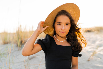 Beautiful brunette   small  girl  in straw hat  posing om the beach. Sunset warm colors.