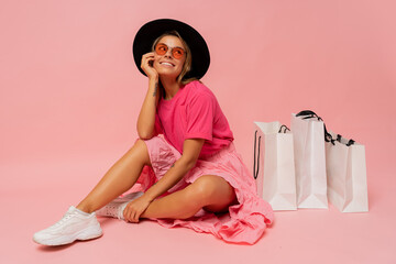 Pretty smiling woman in  spring trendy outfit posing with white shopping bags in studio over pink...