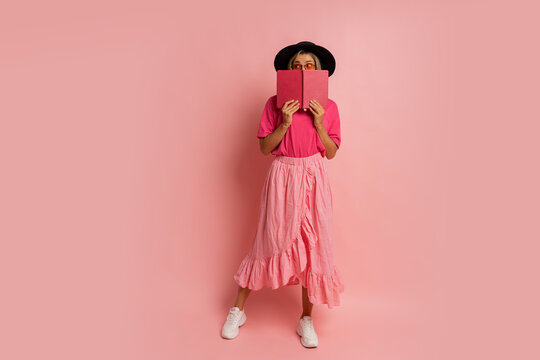 Cute blond woman in black hat and pink spring outfit holding book and  posing in studio.  Full lenght.