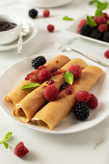 Rolled blini with jam, fresh raspberries and blackberries decorated with mint on a white plate for breakfast or lunch