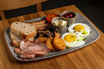 A plate with big english breakfast: fried eggs, hash browns, bacon, mushrooms, sausages, grilled tomato, bread, butter, cherry jam
