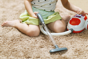A small girl is playing and cleaning the room with a toy vacuum cleaner, sitting on the carpet.A cute little girl cleans herself, vacuums the carpet in the room at home.Children's games, entertainment