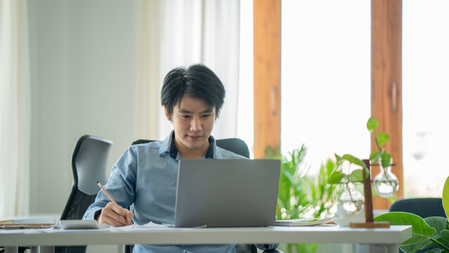 Young asian businessman smiling while working with laptop computer at office, business office lifestyle concept