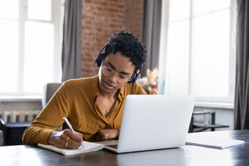 Focused Black student girl in glasses and headphones writing notes at laptop, checking handwriting summary, watching webinar, listening learning audio lecture, taking online virtual training course
