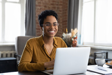 Joyful happy short haired Black business woman in glasses sitting at workplace with laptop in home office, looking at camera, smiling. Millennial worker, employee, entrepreneur head shot portrait