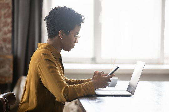 Concentrated African Gadget User Woman Using Online App, Virtual Bank Service On Mobile Phone, Typing Text Message, Sitting At Table, Working At Laptop Computer In Home Office. Side View