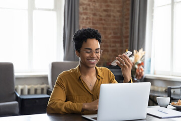 Happy confident African freelance professional woman talking on speaker on cellphone at home...