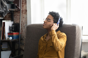 Calm inspired millennial Black girl wearing glasses, headphones, listening to music, resting in...