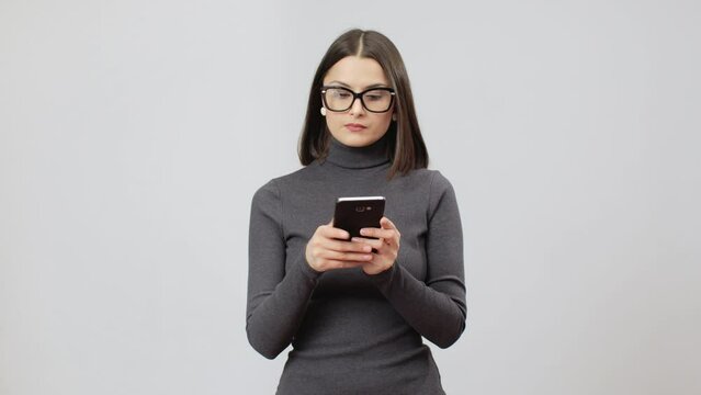 Caucasian woman in glasses and gray turtle neck dress on a grey background serfing social networks on her cell phone