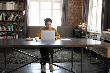 Focused Black business woman in home office full length candid portrait. Serious African freelance worker, entrepreneur, student using laptop at wide wooden table workplace with coffee cup and snacks © fizkes