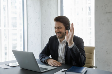 Happy young handsome businessman in suit wearing wireless headphones waving hand, looking at laptop screen, starting video call conversation with colleagues or communicating distantly with clients.