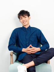 Portrait of handsome Chinese young man with curly black hair in blue shirt sitting in armchair and posing against white wall background. Fingers crossed and smiling at camera confidently, front view.