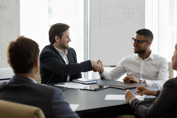 Happy young Caucasian businessman shaking hands with African American partner, making deal at brainstorming meeting with diverse colleagues, sitting at table in modern office, cooperation concept.