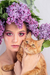 Portrait of a young girl in the studio with lilac flowers on her head and a red kitten in her arms. Beautiful doll-style make-up with lilac flowers on her head and a red kitten in her hand.