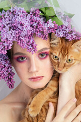 Portrait of a young girl in the studio with lilac flowers on her head and a red kitten in her arms. Beautiful doll-style make-up with lilac flowers on her head and a red kitten in her hand.