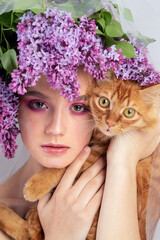 Portrait of a young girl in the studio with lilac flowers on her head and a red kitten in her arms. Beautiful doll-style make-up with lilac flowers on her head and a red kitten in her hand.