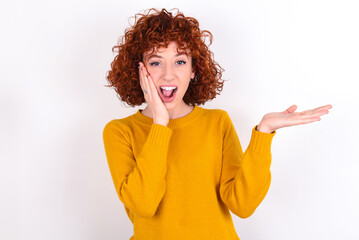 Positive glad young redhead girl wearing yellow sweater over white background says: wow how exciting it is, indicates something.  One hand on his head and pointing with other hand.