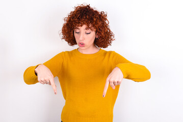 Amazed young redhead girl wearing yellow sweater over white background points down with fore...