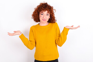 Puzzled and clueless young redhead girl wearing yellow sweater over white background with arms out,...