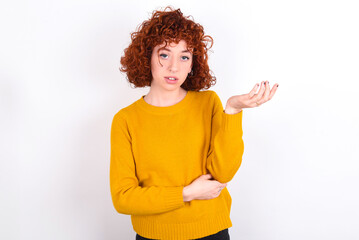 Studio shot of frustrated young redhead girl wearing yellow sweater over white background gesturing with raised palm, frowning, being displeased and confused with dumb question.